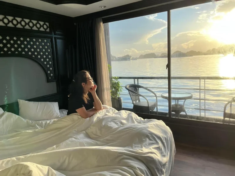 Woman enjoying panoramic view of Lan Ha Bay from bed in luxury cruise cabin with private balcony - Doris Cruise 2 days 1 night